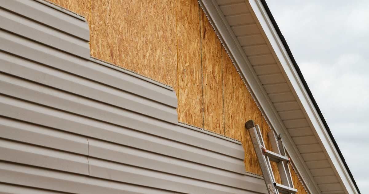 A house under renovation with partially installed beige vinyl siding, exposing the wooden sheathing underneath. A ladder is leaning against the wall.